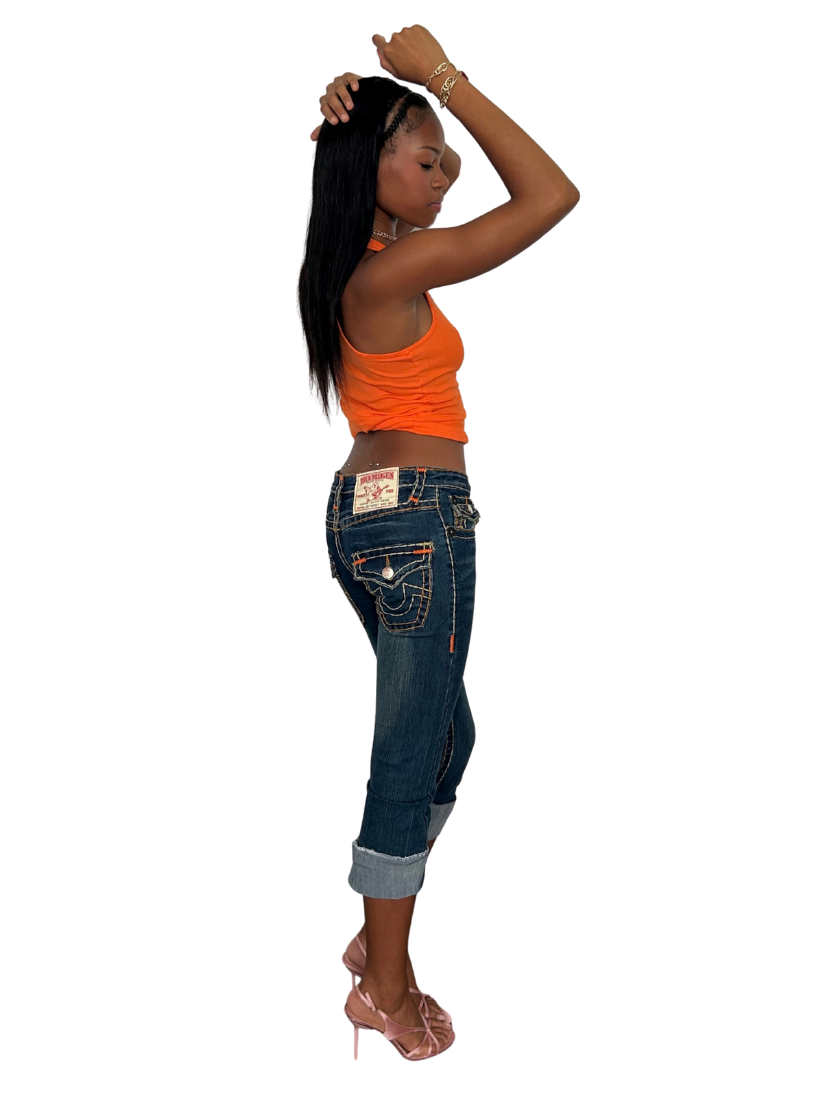 A woman posing in a side profile view, wearing a bright orange ribbed tank top, dark blue denim jeans with rolled cuffs, and pink high-heeled sandals, set against a solid white background.