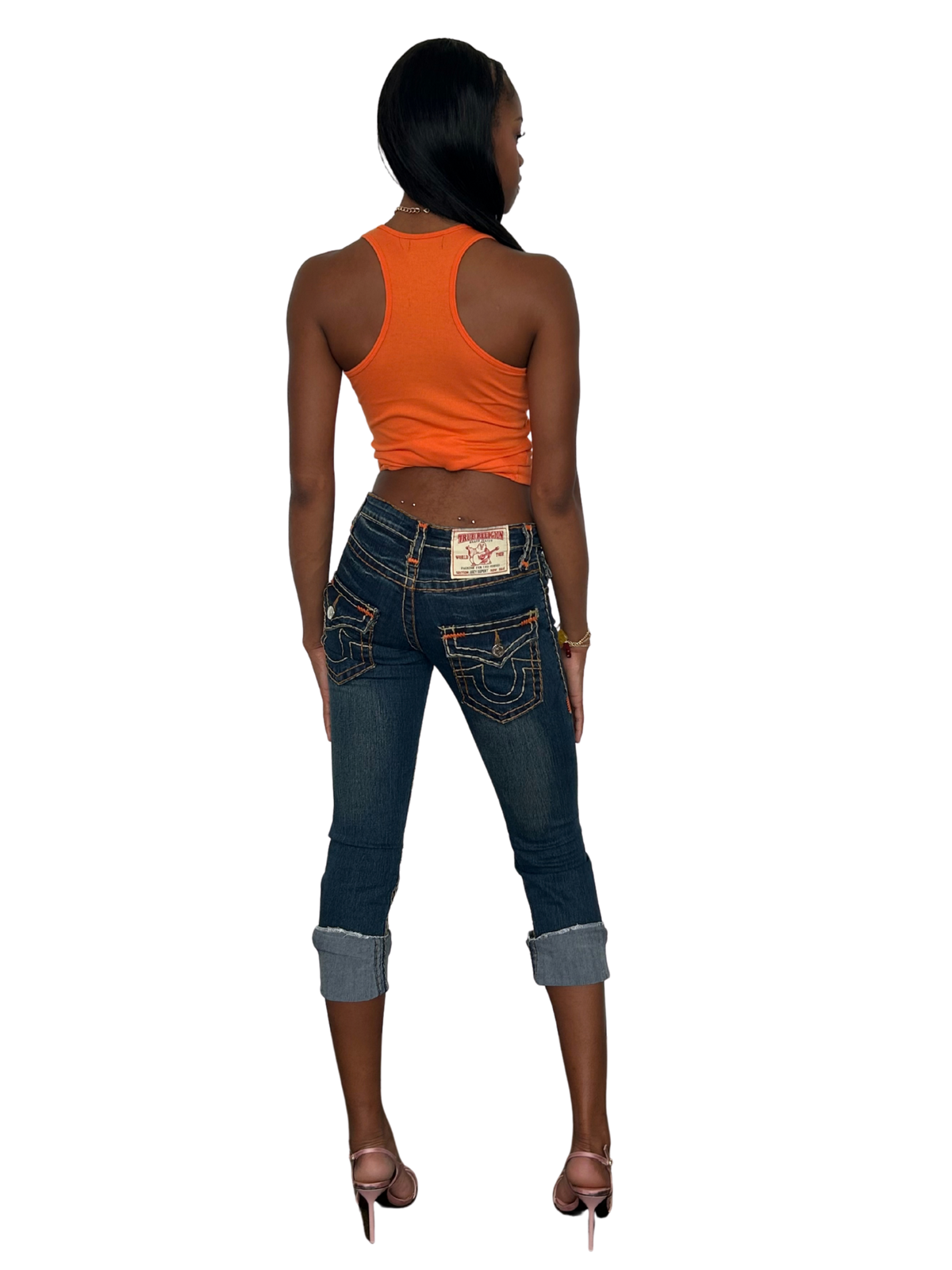 A woman shown from the back wearing a bright orange ribbed tank top, dark blue denim jeans with rolled cuffs, and pink high-heeled sandals against a plain white background.