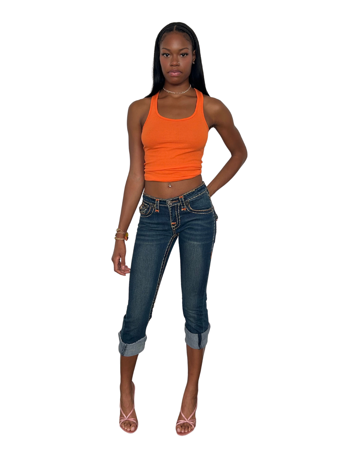 A woman modeling a bright orange ribbed tank top and dark blue cropped denim jeans with rolled cuffs, paired with strappy high-heeled sandals, set against a plain white background.