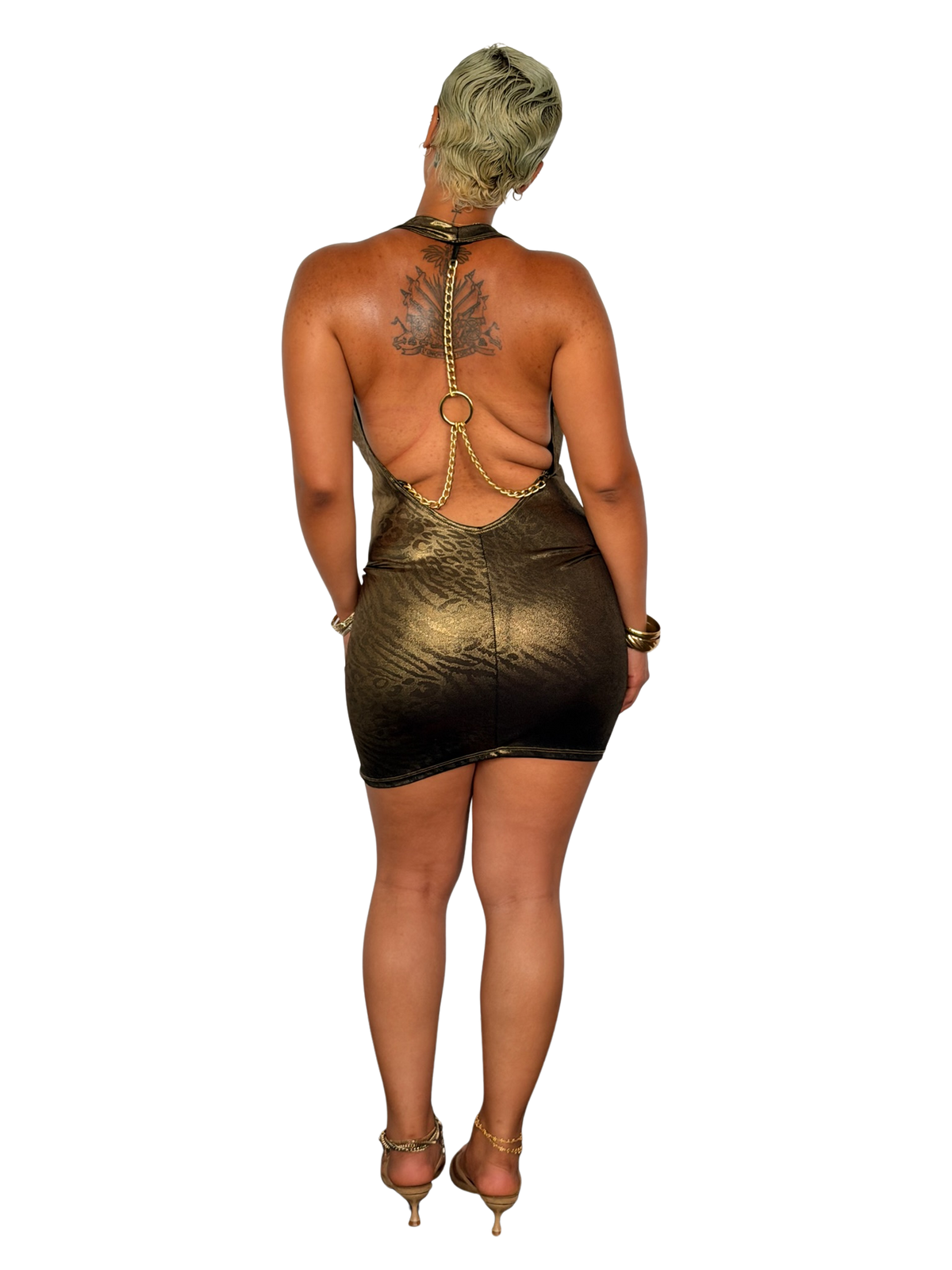 A woman shown from the back wearing a metallic gold mini dress with an open back and chain detailing, paired with gold high-heeled sandals, against a plain white background.