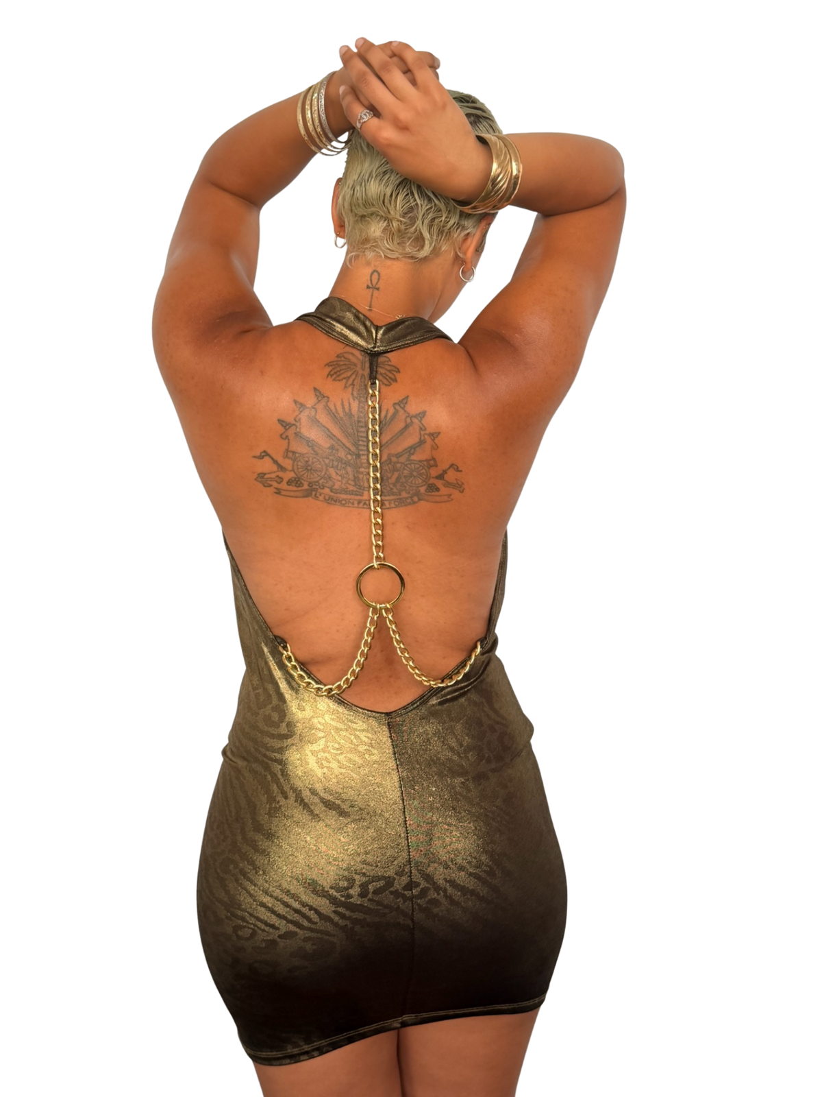 A woman shown from the back wearing a metallic gold open-back mini dress with chain detailing and a circular ring at the center, photographed against a plain white background.