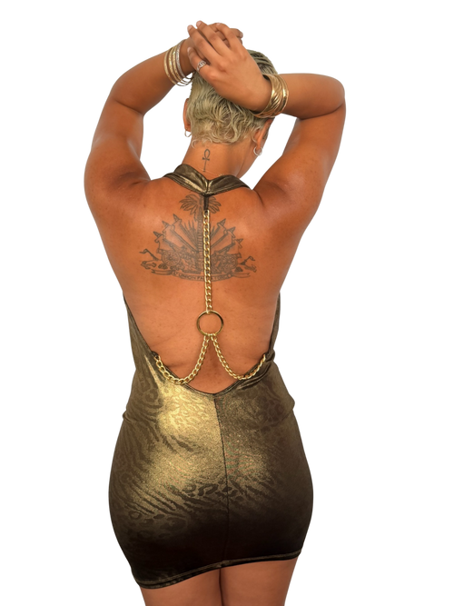 A woman shown from the back wearing a metallic gold open-back mini dress with chain detailing and a circular ring at the center, photographed against a plain white background.